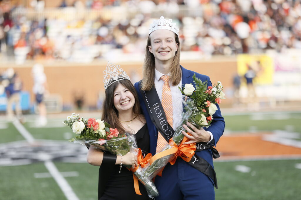 Two students wearing crowns and sashes stand on a football field holding bouquets, smiling at the camera.