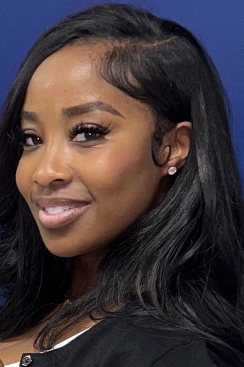 Woman with long black hair, wearing a black cardigan and smartwatch, smiling in front of a plain blue background.