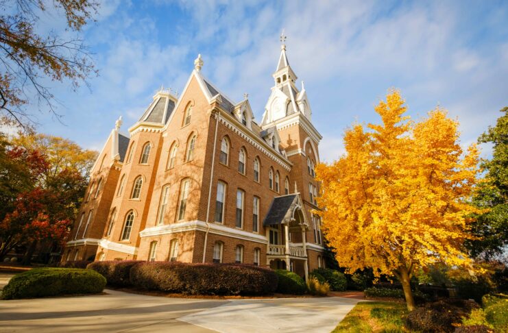 Historic brick building with pointed towers surrounded by autumn trees under a partly cloudy sky.