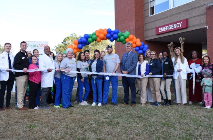 A group of people stand in front of an emergency room entrance, holding a ribbon for a ribbon-cutting ceremony.