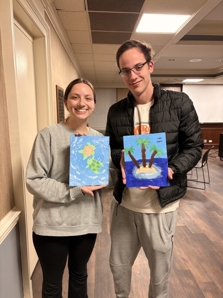 Two people standing indoors, each holding a small canvas painting featuring tropical island scenes.