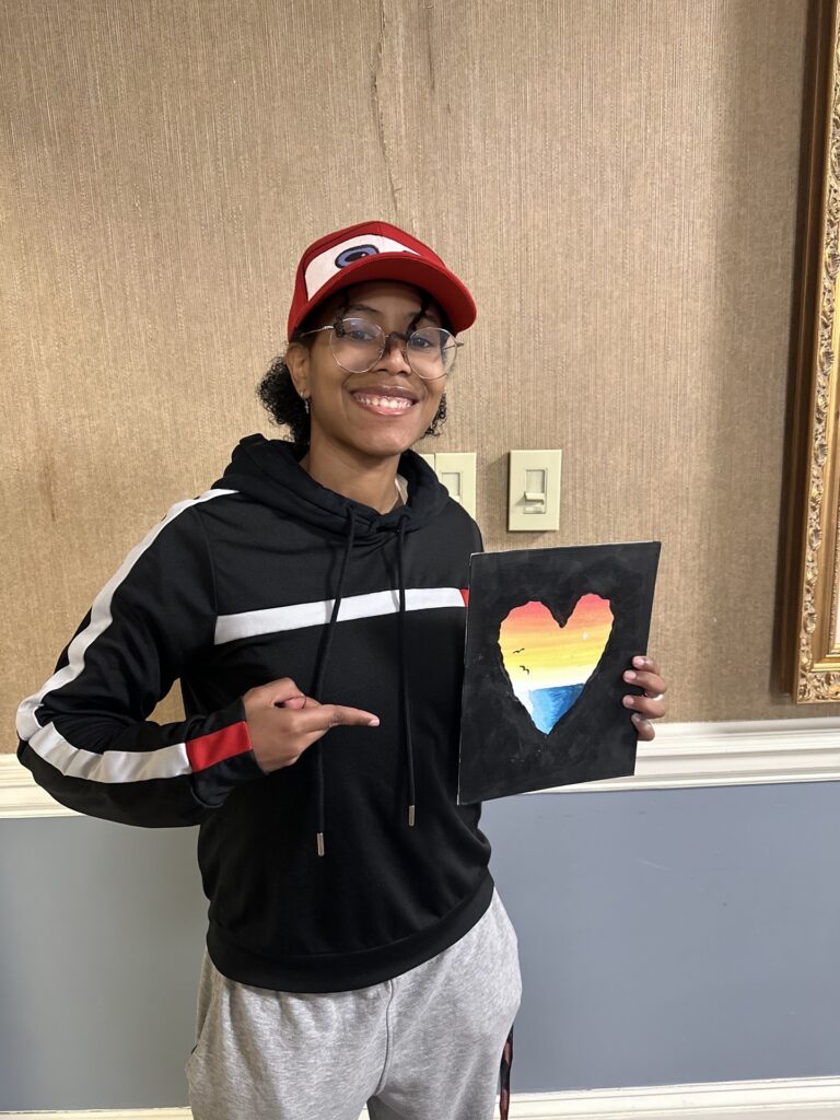 Person in a black hoodie and red cap smiles and points at a heart-shaped painting with rainbow colors.