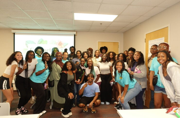 A group of people pose together and smile in a classroom with a presentation projected on the wall behind them.