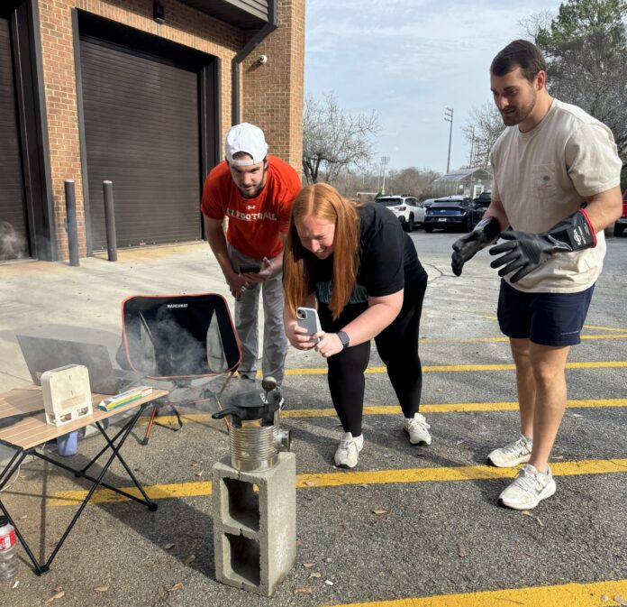 Three people observe and record an experiment with a smoking container on cinder blocks in a parking lot.