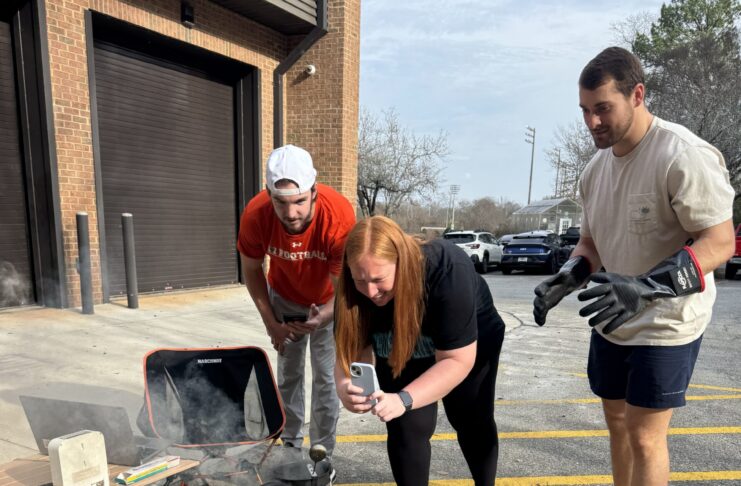 Three people observe and record an experiment with a smoking container on cinder blocks in a parking lot.