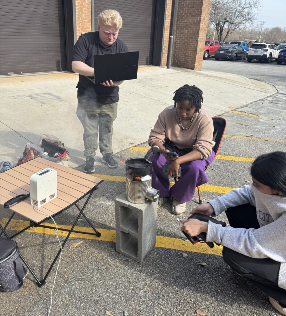 Three people gather around a small outdoor stove on cinder blocks; one holds a laptop, another tends to the stove.