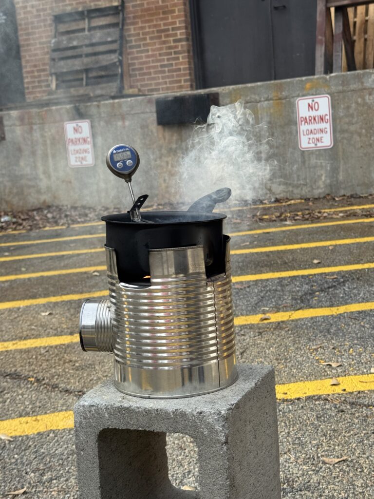 A metal can stove with a thermometer and smoking pot sits on a cinder block in a parking lot near No Parking signs.