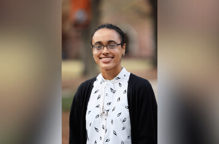 A woman wearing glasses, a white blouse with a black pattern, and a black cardigan stands outdoors, smiling.