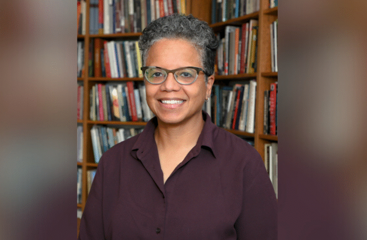 A person with short curly hair and glasses smiles while standing in front of a bookshelf filled with books.