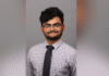 Junior selected for highly competitive Hertog Fellowship A person wearing glasses, a checkered shirt, and a black tie poses in front of a plain gray background.