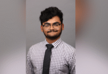 Junior selected for highly competitive Hertog Fellowship A person wearing glasses, a checkered shirt, and a black tie poses in front of a plain gray background.