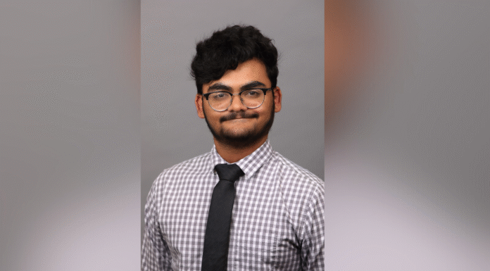 Junior selected for highly competitive Hertog Fellowship A person wearing glasses, a checkered shirt, and a black tie poses in front of a plain gray background.