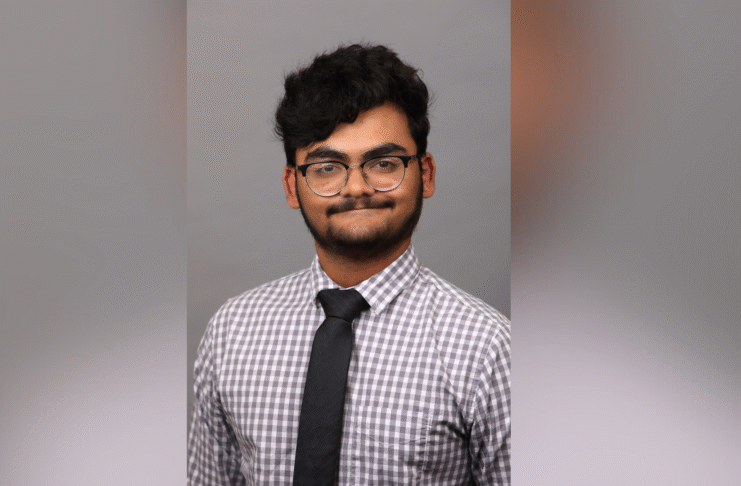 A person wearing glasses, a checkered shirt, and a black tie poses in front of a plain gray background.