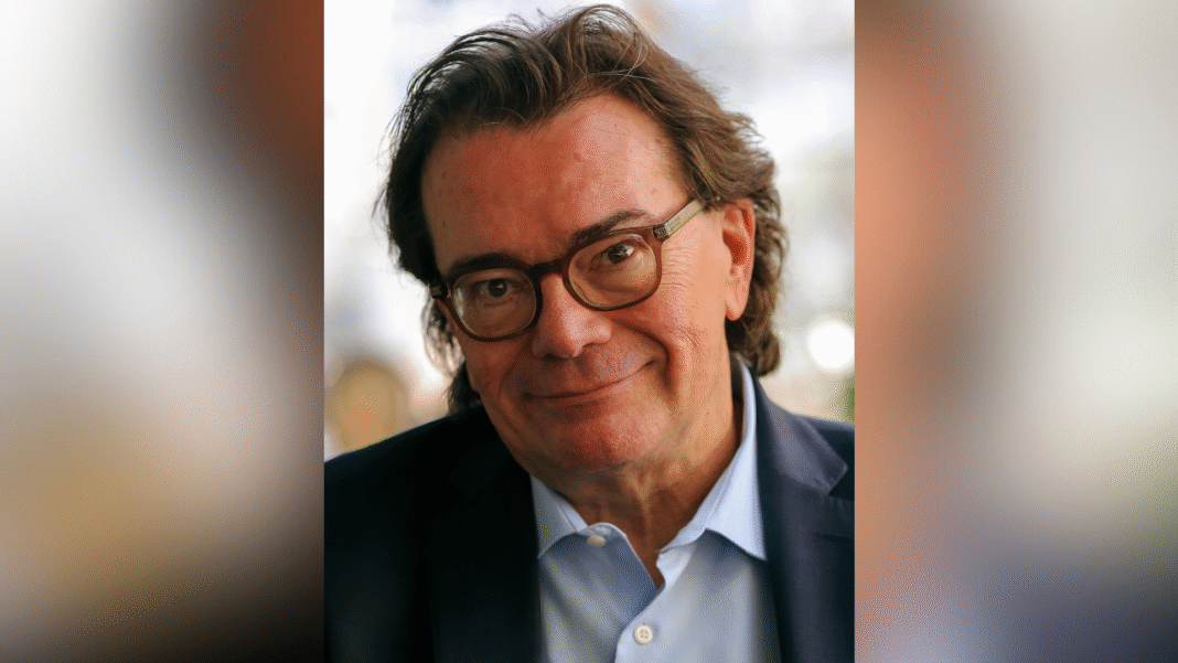 A middle-aged man with glasses and dark hair, wearing a suit jacket and light blue shirt, smiles at the camera.