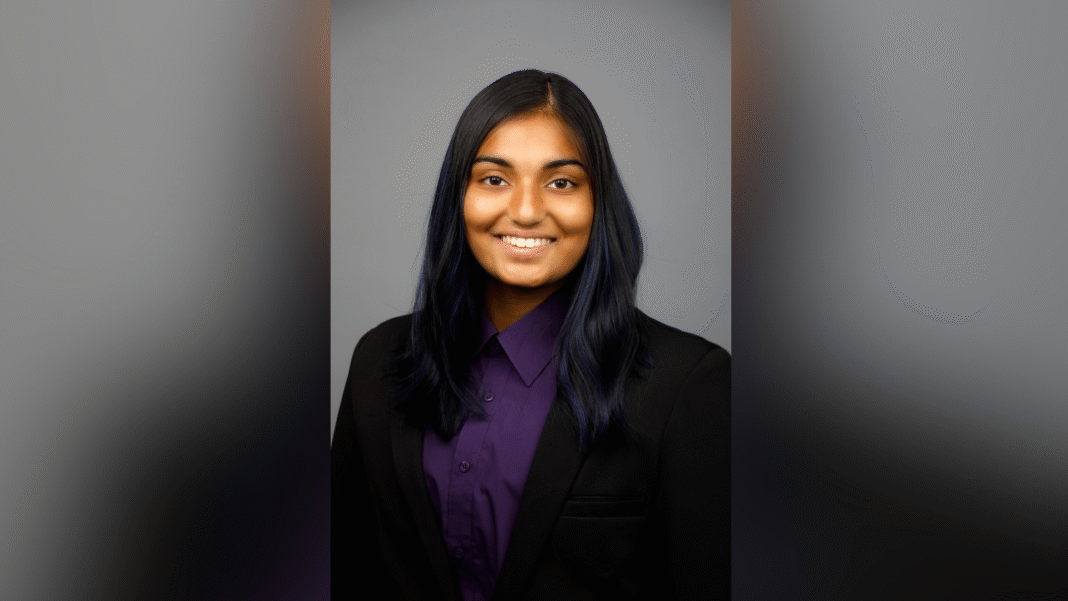 A person with long dark hair, wearing a black blazer and purple shirt, smiles at the camera against a gray background.