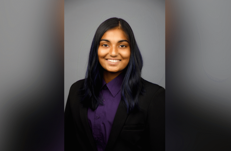 A person with long dark hair, wearing a black blazer and purple shirt, smiles at the camera against a gray background.