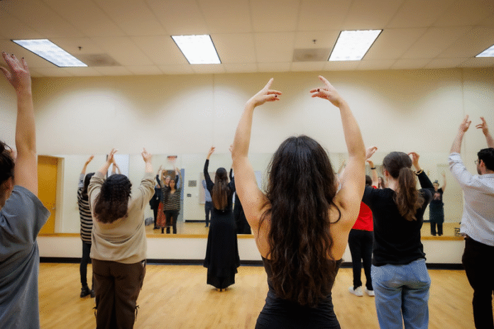 A group of people practice dance in a mirrored studio, raising their arms in unison.