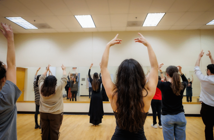 A group of people practice dance in a mirrored studio, raising their arms in unison.