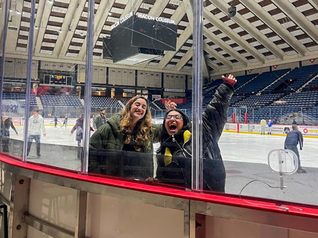 Two people smile and pose by the rink glass at an indoor ice skating arena with others skating in the background.