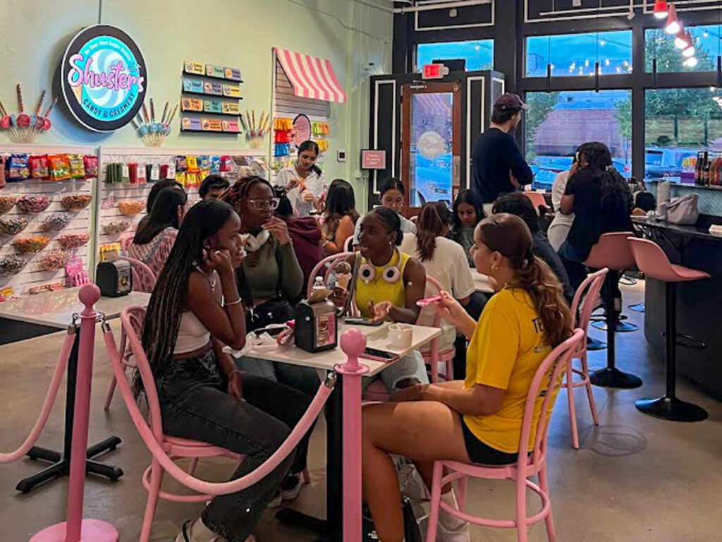 Group of people sitting at pink tables in a brightly lit candy shop with colorful sweets on display shelves.