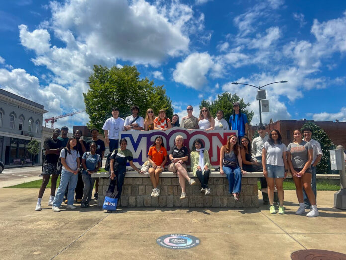 A group of people pose together outdoors in front of a large Macon sign on a sunny day.