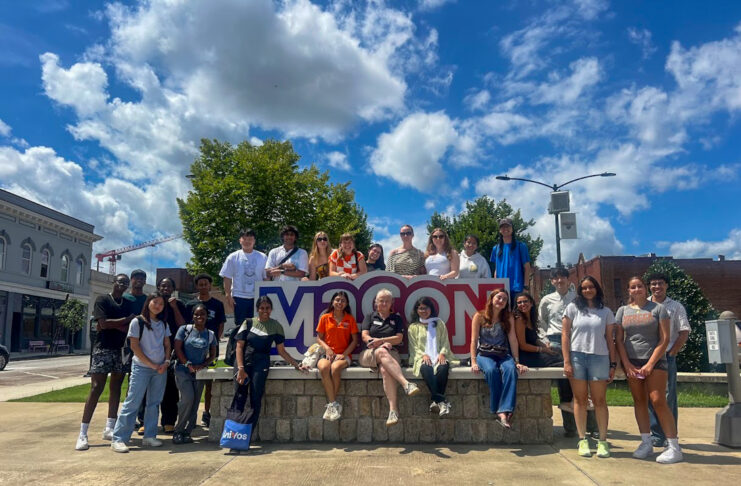 A group of people pose together outdoors in front of a large Macon sign on a sunny day.
