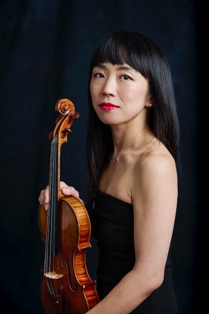 Woman in a black dress holding a violin against a dark background, looking toward the camera.
