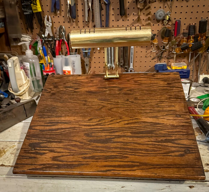 A wooden desktop stand with a brass lamp on top, set on a workbench with various tools hanging in the background.