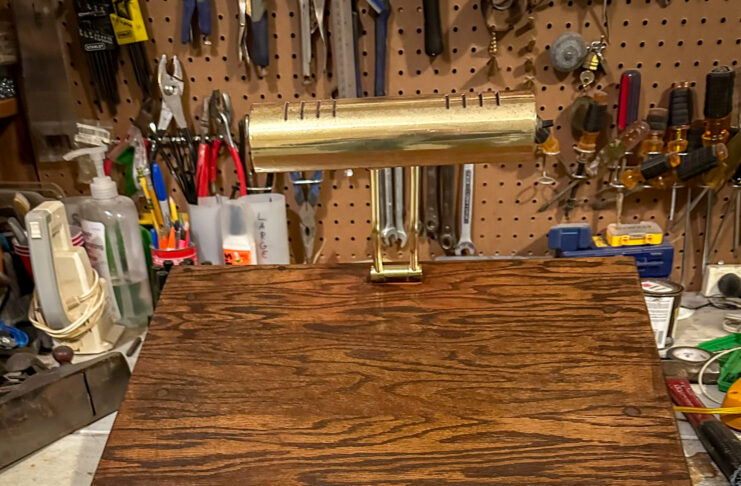 A wooden desktop stand with a brass lamp on top, set on a workbench with various tools hanging in the background.