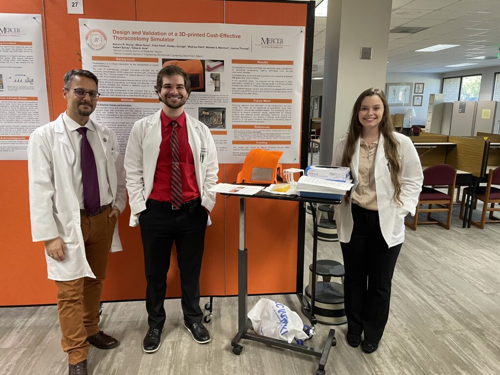 Three people in lab coats stand in front of a research poster and a demonstration table in a library or office setting.