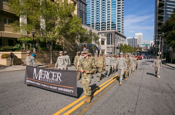A group of people in military uniforms march on a city street holding a Mercer University banner.