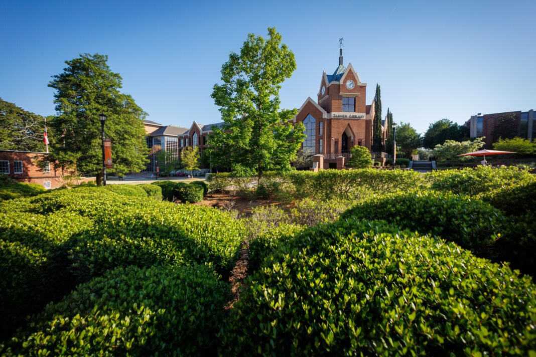 Red brick university building with a clock tower, surrounded by green shrubs and trees under a clear blue sky.