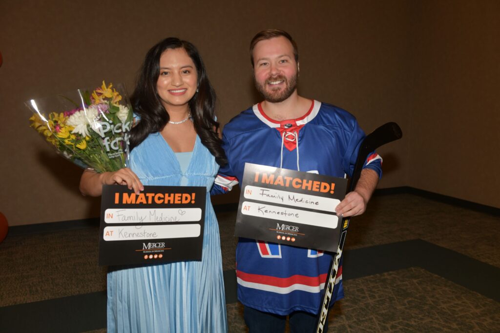 Two people hold signs and smile; one holds flowers, both matched for Family Medicine at Kennestone, Mercer University.