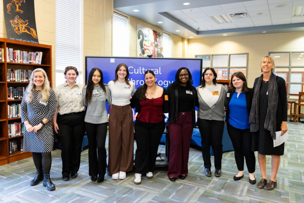 Nine people stand in a row, smiling, in front of a screen displaying the words Cultural Anthropology.