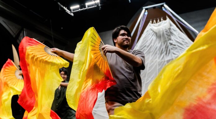 Original Mercer Theatre show brings circus-style spectacles to Historic Quad Two people wave large orange and yellow fabric fans on stage in front of a white, tent-like backdrop.