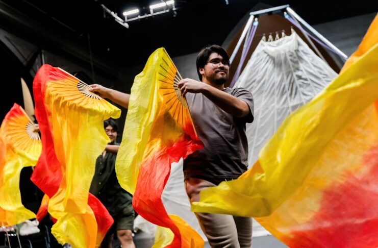 Two people wave large orange and yellow fabric fans on stage in front of a white, tent-like backdrop.