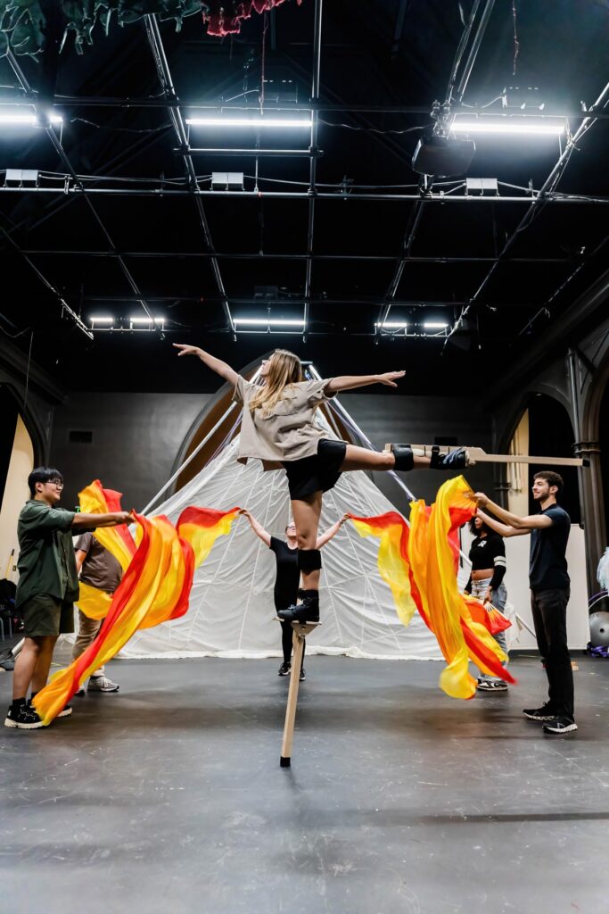 Performer on stilts balances with arms outstretched as others wave large orange and yellow fabric on a stage.