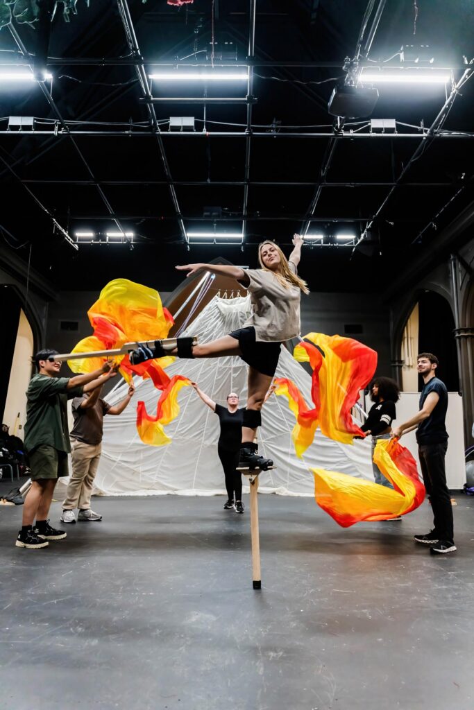 A woman balances on stilts as others wave orange and yellow fabric in a theater rehearsal space.