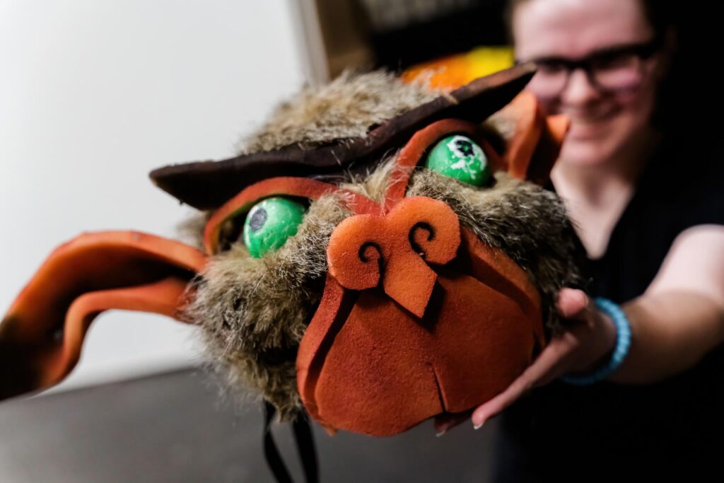 Person holding a large, furry, handmade animal mask with green eyes and exaggerated facial features.
