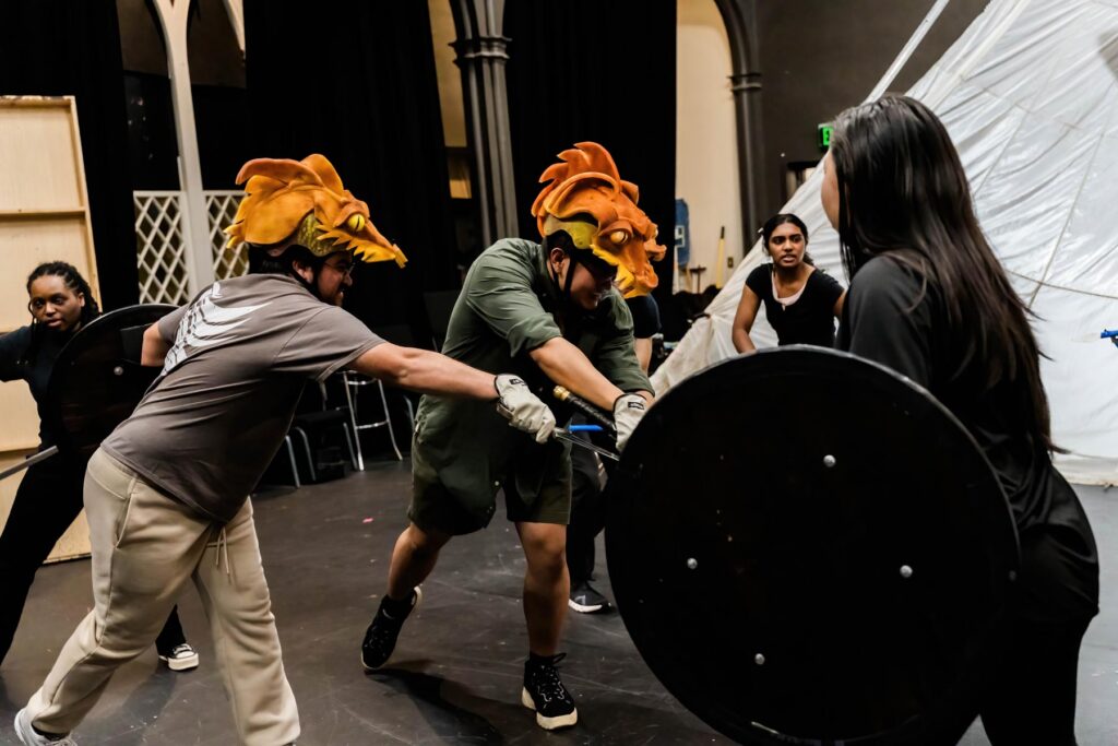 Two people wearing dragon headpieces practice stage combat with swords and shields as others watch in a theater setting.