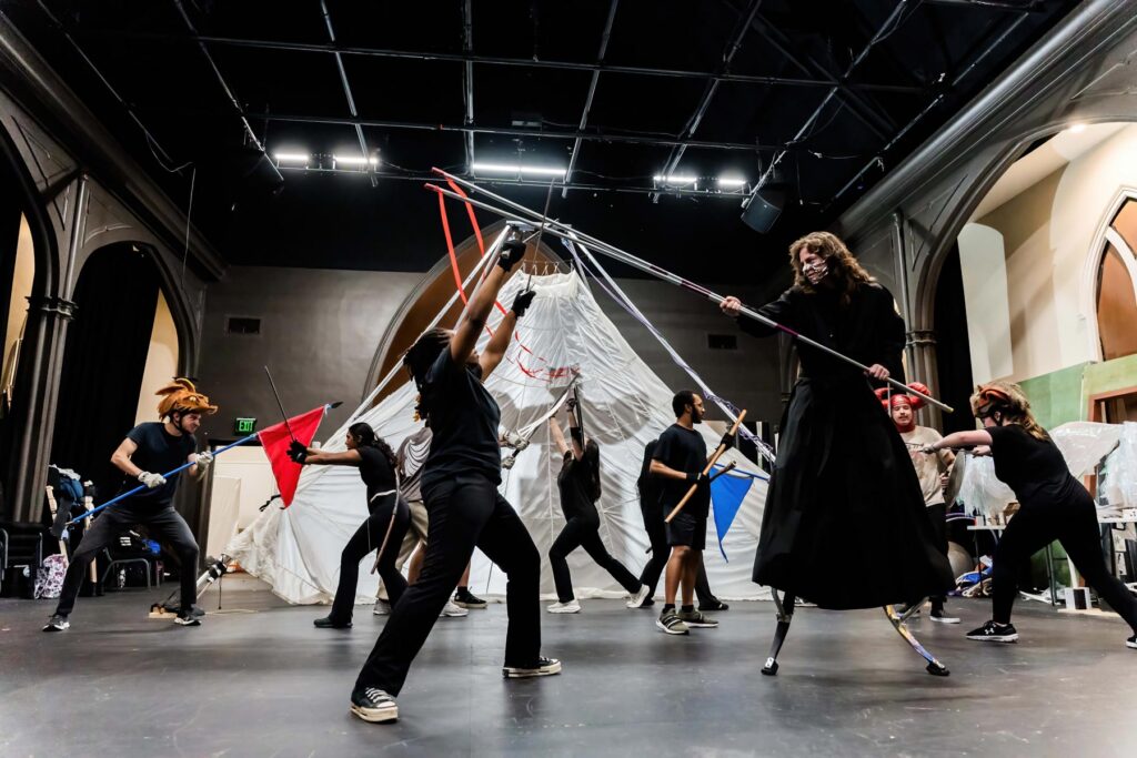 A group of people in costume rehearses a dramatic scene with props on a stage in a theater setting.
