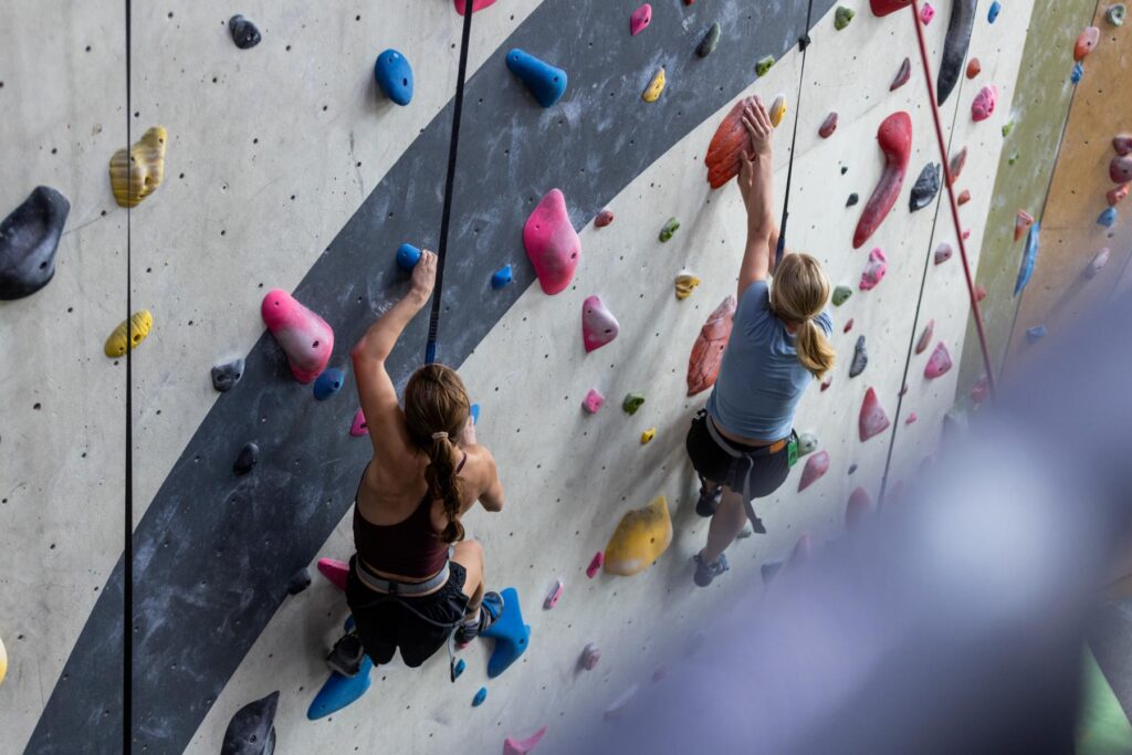 Two people climb an indoor rock climbing wall with multicolored holds and safety harnesses.