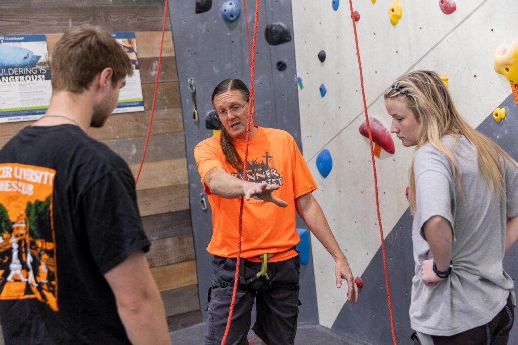 An instructor explains climbing safety to two people at an indoor rock climbing gym.