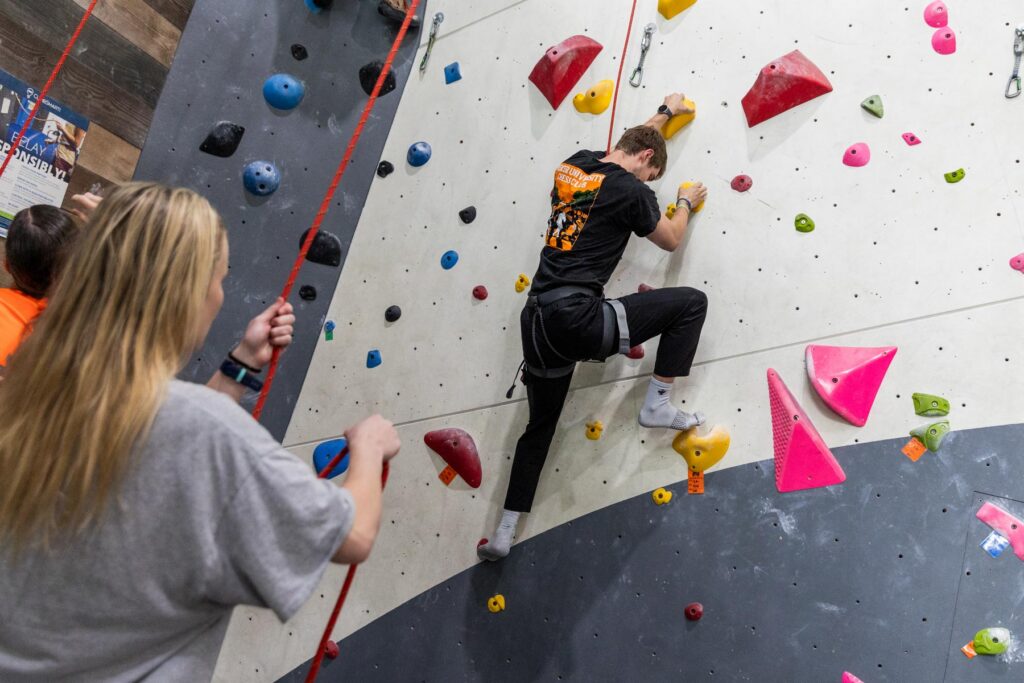 A person climbs an indoor rock wall while another person belays and watches from below.