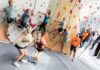 Students reach new heights in rock climbing class A group of people climb an indoor rock wall while two women in front smile and gesture thumbs up at the camera.