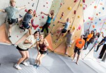 Students reach new heights in rock climbing class A group of people climb an indoor rock wall while two women in front smile and gesture thumbs up at the camera.