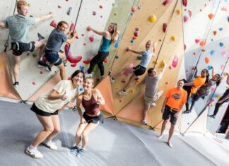 Students reach new heights in rock climbing class A group of people climb an indoor rock wall while two women in front smile and gesture thumbs up at the camera.