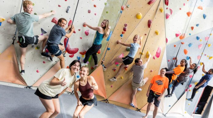Students reach new heights in rock climbing class A group of people climb an indoor rock wall while two women in front smile and gesture thumbs up at the camera.
