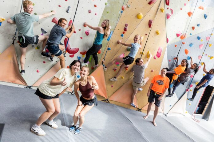 A group of people climb an indoor rock wall while two women in front smile and gesture thumbs up at the camera.