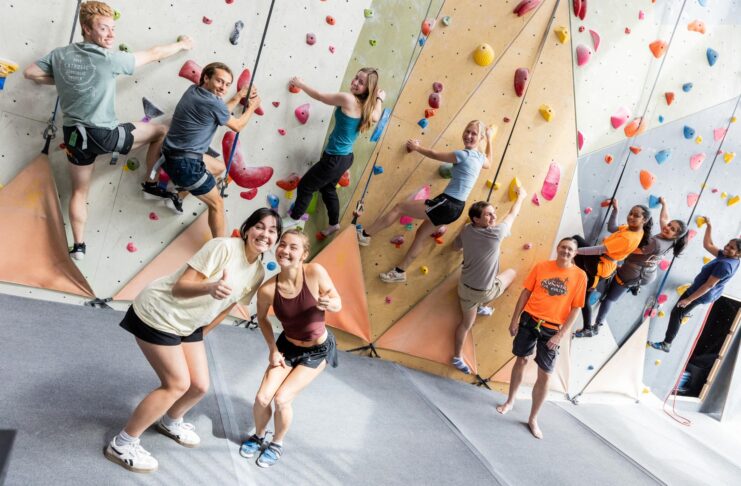 A group of people climb an indoor rock wall while two women in front smile and gesture thumbs up at the camera.